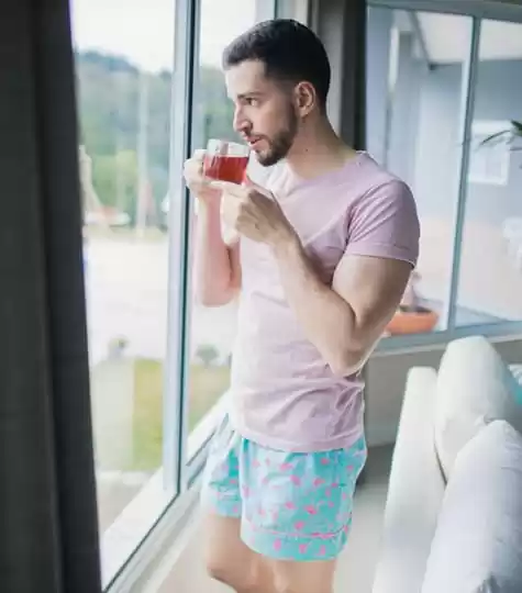 Homem tomando chá de pé enquanto olha pela janela, usando o shorts masculino do pijama Naps na estampa Flamingoland