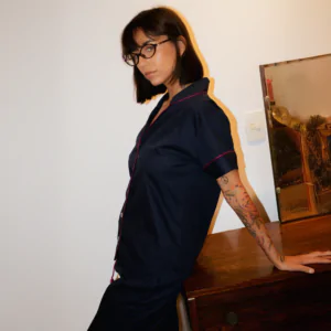 Mulher usando pijama azul marinho de manga curta e calça, com acabamento bordô, em tecido 100% algodão, apoiada em um móvel de madeira.