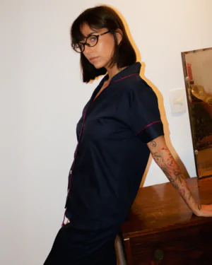Mulher usando pijama azul marinho de manga curta e calça, com acabamento bordô, em tecido 100% algodão, apoiada em um móvel de madeira.